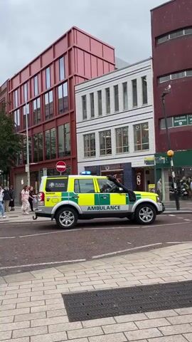 The video opens on a street scene with several buildings lining the right side. A yellow and green ambulance is parked on the right side of the street, facing away from the camera. The ambulance has "AMBULANCE" written on its side. To the left of the ambulance is a row of shops. In the foreground, there is a pedestrian crossing and a textured pavement. On the left side of the street, there is a red building and a white building with multiple windows. A few people are visible walking on the left side of the street, one pushing a stroller. The sky appears overcast.
