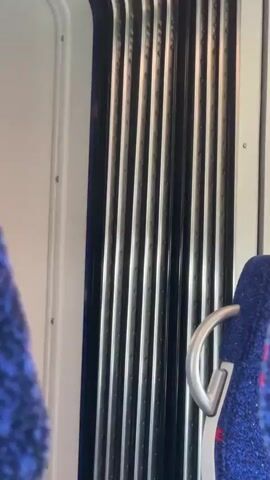 The video shows the interior of a train, with a focus on a section of the wall and a seat. The wall is white with visible screws or rivets and has a vertical, ribbed, black and silver panel. A blue fabric seat with a silver grab bar is partially visible on the right side of the frame. Another similar seat is partially visible on the left. The train appears to be moving, indicated by the subtle vibrations and sounds.