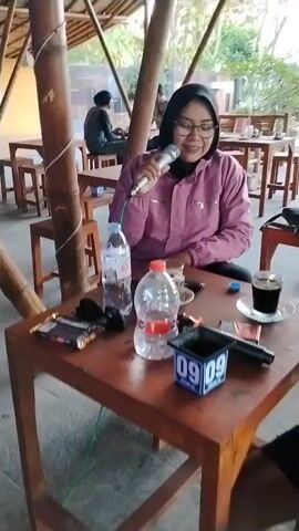 A person wearing glasses and a headscarf, dressed in a pink jacket, holds a microphone to their mouth while sitting at a wooden table. Their hands are covering their mouth as they sing or speak into the microphone. On the table are two plastic water bottles, a cup of coffee, a chocolate bar, cigarettes, and a small container with the numbers "09" on it. In the background, other people are seated at tables, and the environment appears to be an outdoor or semi-outdoor seating area with wooden structures and greenery.