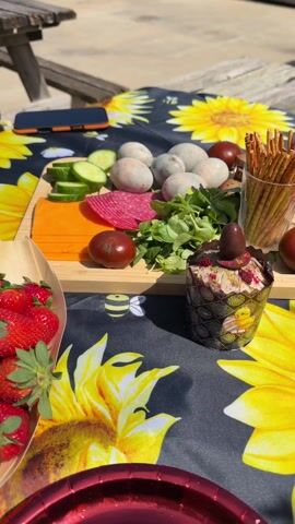 The video shows a table set for a picnic, with a sunflower-themed tablecloth. Visible on the table are a wooden tray with strawberries, a cutting board with cheese, cucumber slices, salami, tomatoes, and lettuce, a heart-shaped box with painted Easter eggs, a can of beer, a white vase with flowers, a decorative heart-shaped frame, and red paper plates. A person is seen adjusting the items on the table. The scene appears to be outdoors in bright sunlight.