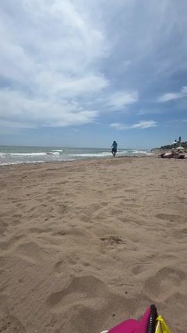 The video shows a sandy beach with the ocean in the background under a cloudy sky. In the first frame, a person is standing near the water's edge, facing the sea. In the second frame, this person appears to be running away from the water. The subsequent frames show the person moving further into the distance, becoming a smaller figure in the frame as they approach the water's edge once more. The camera angle is low, close to the sand, and angled upwards towards the sky and the horizon. There are other people visible in the far distance on the beach, and some objects, possibly beach towels, are visible in the foreground, suggesting a recreational setting.
