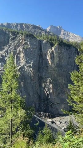 The video shows a close-up view of a narrow waterfall cascading down a sheer, gray rock face. The waterfall is thin and appears to be a series of trickles rather than a powerful torrent. The rock face is textured and shows various horizontal lines and crevices. Green trees are visible on top of the cliff and in the foreground, with a clear blue sky above. The bottom of the waterfall appears to lead to a rocky area. There are no people or any other discernible objects or actions in the video. The overall impression is a remote, natural mountainous landscape.