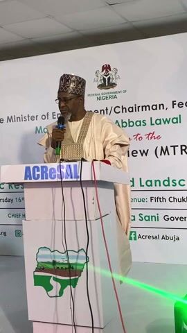 A man in traditional Nigerian garb is speaking at a podium. He is holding a microphone. The podium has a logo for ACRESAL. Behind him is a backdrop with the Federal Government of Nigeria logo. The backdrop also has text about the event. There is green lighting in the foreground.