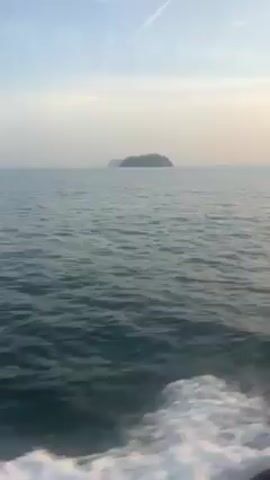 The video shows a view from a boat on the ocean. The water is blue-green and slightly choppy. In the distance, a small, green island is visible on the horizon. The sky is a hazy blue with a faint contrail. The boat is moving, as evidenced by the wake in the foreground.