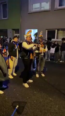 The video shows a group of people standing outdoors, dressed in what appears to be festive attire. Two individuals in the center are wearing cone-shaped hats and scarves with possible team logos. They are holding drinks, raising them in a toast, and one appears to be taking a drink. The rest of the group is also wearing similar scarves. They are standing on a paved surface, possibly a street or sidewalk, flanked by buildings on both sides. There is a blue hose or cable visible on the ground. The scene appears to be taking place at night, with some artificial lighting present. There are other people standing and watching in the background.