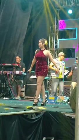 A female performer in a sparkling red dress and high heels dances on a stage. She holds a microphone and moves her arms and body to the music. Behind her, a keyboardist plays an electronic keyboard. Another man stands to her right, also holding a microphone. The stage is lit with colorful spotlights, and a large screen is visible in the background. The scene appears to be a live performance at an outdoor event.
