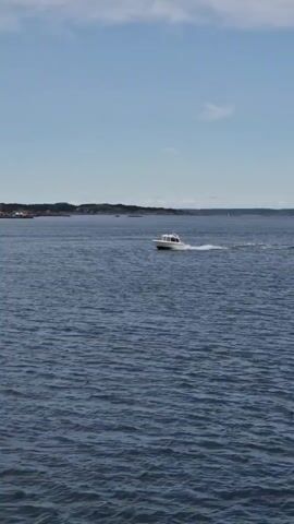 The media file captures a serene maritime scene on a clear day in Öckerö, Sweden. The primary subject is a white motorboat navigating through deep blue water, leaving a white wake behind it. The boat appears to be moving at a moderate speed. In the background, the coastline of Öckerö is visible, characterized by a gently rolling landscape dotted with structures, including a prominent yellow building that stands out against the natural scenery. The sky above is a bright blue, with a few wispy clouds suggesting fair weather. The overall atmosphere is peaceful and suggests a pleasant day for boating. The scene appears to be set during daylight hours, likely in the late morning or afternoon, given the bright illumination. There is no visible text in the media file.