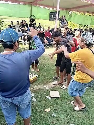 The video shows a group of people dancing outdoors under a tent. The ground is grassy, and there are bottles of what appear to be beer on the ground. Most of the dancers are men, dressed in casual clothing. There are also spectators in the background, some of whom are seated and others standing. A person is standing on a platform behind the dancing crowd, possibly filming the event. There's music playing, and the general atmosphere seems celebratory.