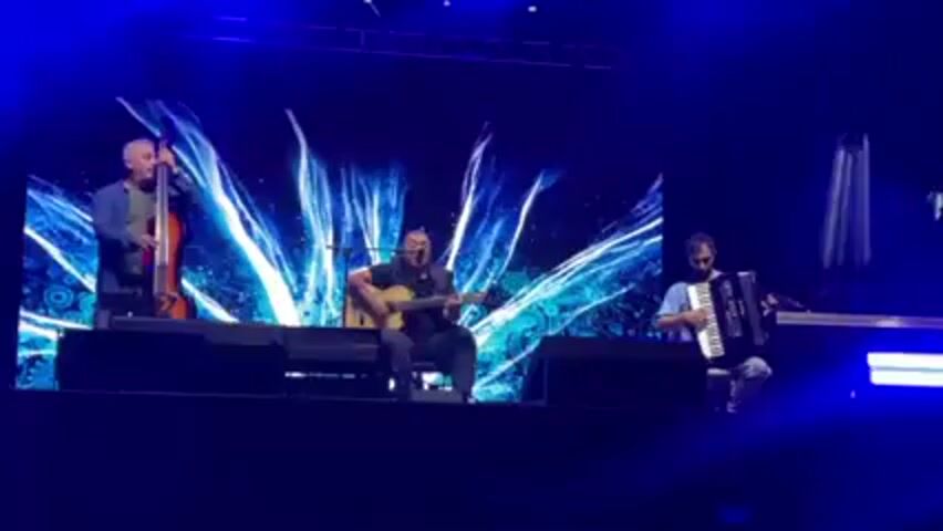Three men are on a stage illuminated by blue lights. The man on the left is playing a stand-up bass. The man in the center is seated and playing an acoustic guitar while singing. The man on the right is seated and playing an accordion. A large screen behind them displays a swirling blue and white pattern.