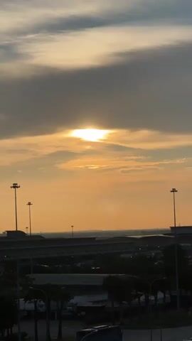 This media file captures a panoramic view of an airport setting during what appears to be sunset. The sun is visible, partially obscured by clouds, casting a warm, golden-orange glow across the sky. The clouds are wispy and streaked, with some darker, heavier clouds towards the top of the frame.

The scene is dominated by the vast expanse of the airport. In the middle ground, a long, low-slung building, likely an airport terminal or support structure, stretches across the frame. Rows of parked cars are visible in a large parking lot adjacent to this building, suggesting ongoing airport operations. Tall light poles with bright lamps are scattered throughout the airport grounds, some illuminated, indicating the transition from day to evening.

In the foreground, a road runs horizontally, with several trucks and cars on it, including a white box truck with visible branding that is partially obscured but appears to say "NX-TRANS" and a partial logo. Another truck with a yellow cab is also present. A few trees line the roadside, some appearing to be palm trees, which, along with the overall architecture and the presence of an airport, suggests a tropical or sub-tropical location. A tall, slender tower, possibly an air traffic control tower, is visible in the distance to the left of the frame.

The overall mood is tranquil, with the fading sunlight creating a sense of peace. The activity appears to be standard airport operations, with vehicles moving and vehicles parked. The time of day is clearly sunset or late afternoon, given the color of the sky and the illumination of the lights. The location context provided is Sepang, Malaysia, which aligns with the visual cues of a large international airport and tropical vegetation. There are no people clearly visible in the frame.