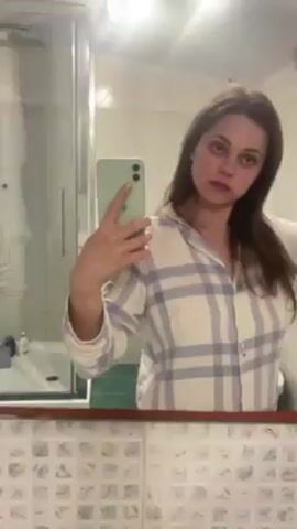 A young woman in a white and blue plaid pajama top stands in a bathroom and takes a selfie in the mirror with a light green smartphone. She is brushing her brown hair. The bathroom has a white shower stall, a mirror with a brown frame, and a tiled wall with a floral pattern.  The lighting in the bathroom is bright.