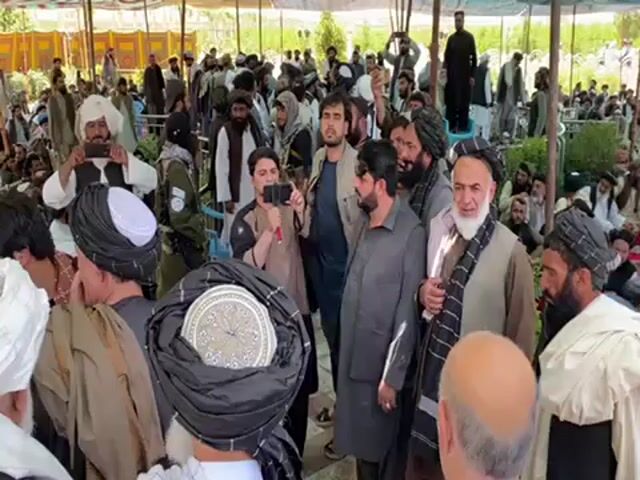 Here is a detailed description of the video:

The video shows a gathering of a large group of men, likely outdoors, under what appears to be a tent or canopy structure that provides shade. The majority of the men are wearing traditional clothing, including turbans, various types of vests, and long robes or shirts. Many have beards and appear to be Middle Eastern or Central Asian in appearance.

Several individuals are holding up their cell phones, seemingly recording or taking photos of something nearby, indicating some point of focus or interest. The men are standing close together, suggesting a crowded environment.

The lighting appears to be bright, indicating a daytime setting. The background includes greenery and glimpses of more people, indicating that the gathering is quite large. There's a generally somber or neutral mood, with no obvious signs of distress or celebration. The scene appears to be a gathering of a cultural or perhaps political nature.