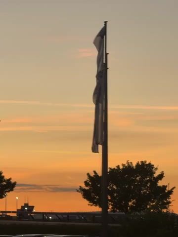 The video shows a flagpole with two flags against a sunset sky. The flags are white with a dark design. In the background, there is a tree and a building that appears to be an airport control tower. The sky is orange and yellow with some faint clouds. The video is shot from a low angle.
