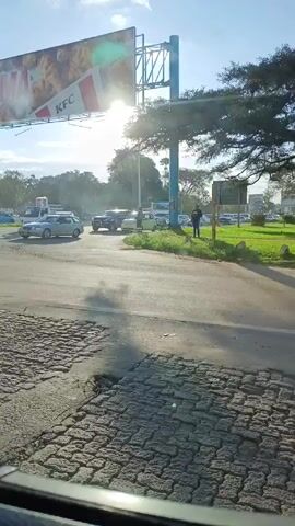 The video shows a view from inside a vehicle, looking out onto a street scene.  A KFC billboard is visible on the left, partially obscuring the sun. Cars are driving and parked along the road, and a person stands on the grassy median. The foreground shows a section of cobblestone pavement. The weather appears sunny with some clouds.