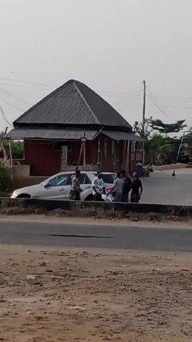 The video shows a roadside scene in what appears to be a rural or developing area. There is a small brick building with a dark grey, peaked roof situated just off the road. A group of five or six people are gathered near a white SUV parked in front of the building. There is a second car further up the road with a third car behind it. Chickens are also visible in the video wandering on the side of the road. The road itself is paved but shows signs of wear and tear, and the surrounding area is dusty and unpaved. Palm trees are scattered in the background.