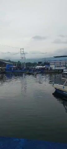 The video shows a waterfront scene with a body of water in the foreground. In the background, there is a blue platform or dock with a tall, white metal structure resembling a crane or tower on it. Various tents, possibly for vendors or events, are set up behind the platform. Many motorcycles are parked along the edge of the platform. Several small boats are moored in the water near the platform. People are visible on the platform, some standing and some appearing to be walking. The sky is overcast, with clouds obscuring the tops of distant hills or mountains. The overall impression is a gathering or event taking place at a waterfront location.