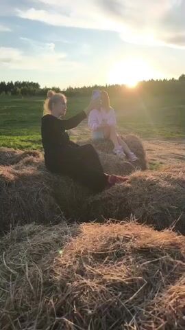 Two young women are seated on large hay bales in an open field during sunset. The sky is bright with a clear horizon and some clouds, and the sun is low, casting a golden light and creating lens flares in the video. A treeline is visible in the background.

The woman in the foreground, on the left, is wearing a black long-sleeved top or dress and black pants, with her hair tied up. She is holding a light-colored smartphone up, appearing to be taking a photo or video, and is smiling towards the camera.

The second woman, further back on the right, is wearing a pink hoodie and light blue shorts with white sneakers. Her hair is down. She is also holding a smartphone and looking down at it, with a subtle smile.

The setting appears rural or agricultural due to the hay bales, with green fields stretching behind them. The overall mood is relaxed and pleasant, capturing a moment during golden hour. No visible text is present in the video.
