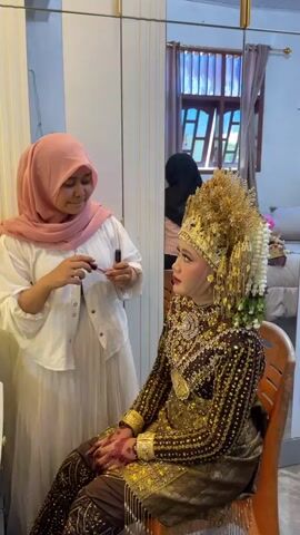 A woman in a pink hijab and a white and cream dress is applying makeup, specifically lipstick, to another woman. The second woman is seated and wearing a traditional, ornate brown and gold wedding outfit, complete with a large, elaborate gold headpiece. Her hands are visible and appear to have henna applied. The setting appears to be an indoor room, possibly a dressing room, with a large mirror in the background. A reflection in the mirror shows another person in a black hijab and a window with wooden blinds. The overall scene depicts a bride being prepared for a wedding ceremony.