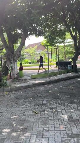 A young person is dribbling a basketball in a paved outdoor area. They are wearing a dark shirt and shorts. There is a smaller child in a red shirt and dark shorts standing near a green barrel to the left of the frame. Trees with green leaves provide shade over the area. In the background, there are buildings and more trees. The ground is covered in interlocking brick pavers. The lighting suggests it is daytime.