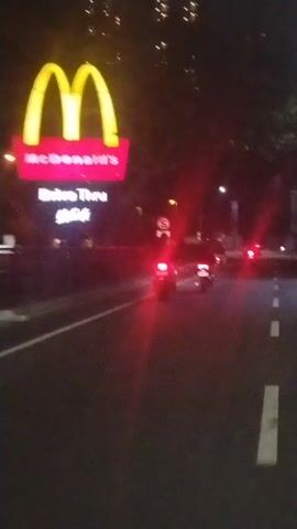 The video is a shaky, first-person perspective of a street at night. On the left side of the screen, a McDonald's sign with the golden arches is visible, lit up in yellow and red. Below it, text reads "McDonald's Drive Thru" and "McCafe". A road with white lane markings is in the foreground. Several motorcycles with illuminated red taillights are moving away from the camera down the road. The environment appears to be urban, with streetlights and building lights visible in the background, as well as trees along the side of the road. The overall lighting is dim, consistent with nighttime.