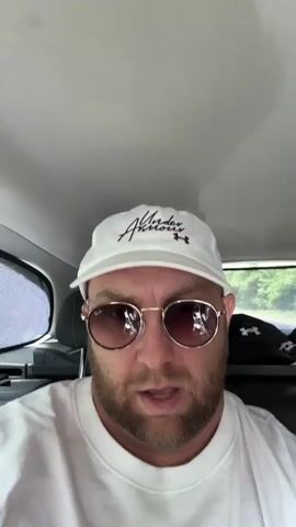 The video is a first-person perspective recording from inside a car. A man with a beard, wearing a white baseball cap and sunglasses, is looking directly at the camera. He is wearing a white sweatshirt. The background shows the interior of the car, including the ceiling and a car window with a sunshade. Outside the car window, trees and another car are visible. The man speaks and smiles at different points in the video.
