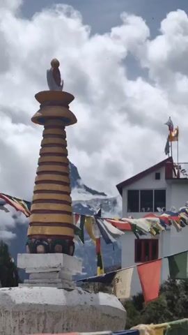 A stupa adorned with yellow and red bands stands prominently in the foreground, its top featuring a figure. Prayer flags in various colors flutter in the breeze, strung across the scene. Behind the stupa, a white building with a red-trimmed roof and red-framed windows is visible. In the distance, a majestic, snow-capped mountain rises against a sky filled with dynamic clouds. The overall atmosphere suggests a serene, spiritual setting in a mountainous region.