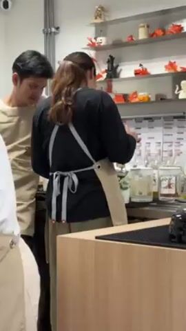 The video shows three people wearing aprons in what appears to be a kitchen or food preparation area. Two are standing behind a counter and working with ingredients, while the third is partially visible at the edge of the frame. The countertop has a shaker, bottles, and other bar or kitchen items, and a shelf behind them displays decorative objects like pumpkins. One of the two individuals at the counter is talking and gesturing with their hands.