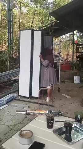 A person wearing a hijab and a grey dress is standing on a stool and adjusting a large, white, foldable screen or partition. The partition is tall and has black trim. The person is on the right side of the partition, pushing it closed with their hands. They are in an outdoor, somewhat cluttered environment. There are metal structures and materials in the background, suggesting a workshop or construction area. In the foreground, there is a table with drinks and other objects. The person appears to be working on setting up or moving the partition.