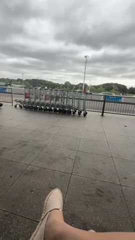 The video begins facing a row of shopping carts on a paved area under a cloudy sky. The carts are lined up to the left. To the right, a metal railing separates the paved area from a road where cars are driving. Beyond the road, there are buildings and trees. The camera pans to the right, revealing more of the road and the surroundings. A person is seen pushing a luggage cart towards the right side of the frame. The camera continues to pan, showing the exterior of a building with a triangular roof structure. Another person is seen walking in the distance. The video ends with a focus on the exterior of the building and the cloudy sky.