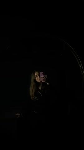 A young woman with long brown hair is taking a selfie in a dark room, likely in front of a mirror. She is wearing a black outfit with white trim and a gold necklace. She is holding a pink phone. The room is almost completely dark, with only a faint light illuminating her face and phone. There is a curved object visible on the right, likely part of the mirror's frame. She smiles slightly while holding up her phone.