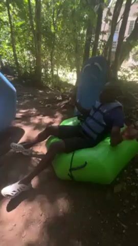 A man, wearing a blue shirt, black shorts, a blue life vest, and white shoes, sits on a green inflatable chair. He is positioned on a dirt ground surrounded by trees, with sunlight filtering through the leaves. There is another blue inflatable object and a paddle board behind him. He is gesturing with his hands. He seems to be talking about the river which he says is moving very fast.