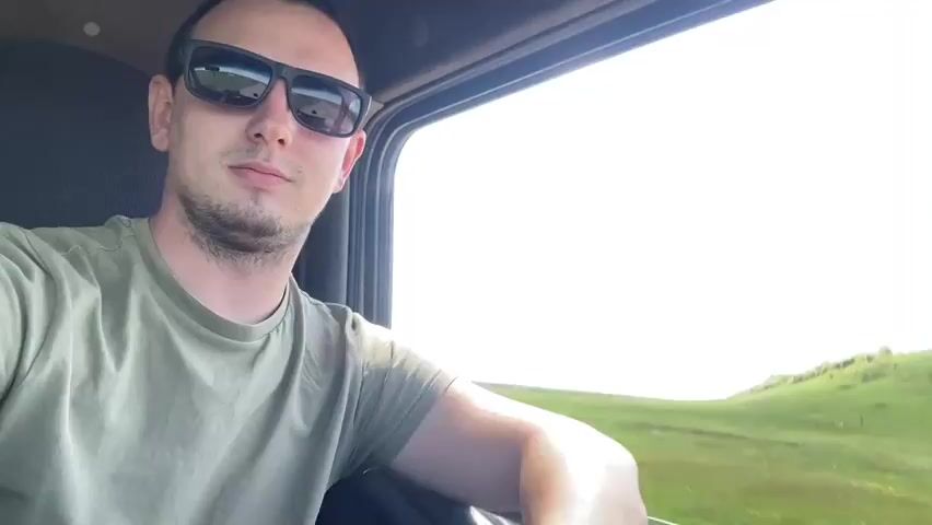 A man wearing sunglasses and a green t-shirt is in the driver's seat of a vehicle. He looks at the camera. The vehicle appears to be moving and the landscape outside the window is green and hilly. A roadside marker painted in the colors of the Russian flag is visible. The camera switches to an exterior view through the windshield. The vehicle is driving on a narrow paved road through a green, grassy landscape under a blue sky with scattered white clouds.