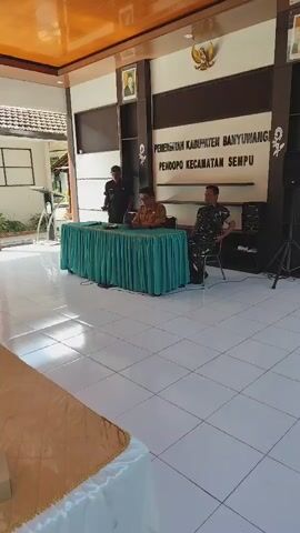 This video shows an indoor event likely taking place at a government building, as indicated by the signage that reads "PEMERINTAH KABUPATEN BANYUWANGI PENDOPO KECAMATAN SEMPU." In the background, three individuals are seated at a table draped with a green cloth. One person, wearing a brown uniform, is speaking into a microphone. To their left, another person in a dark uniform stands, and to their right, a person in a camouflage uniform sits. In the foreground, a long table is covered with a gold cloth, and several people, predominantly women wearing hijabs, are seated on chairs facing the speakers. Some of them appear to be listening, while others are engaged with their phones or looking away. Small food items are visible on small decorative plates placed on the table. The room has a tiled floor and an orange ceiling with lighting fixtures. White decorative trim is visible along the roofline of the building's exterior, seen through an open-air section of the venue.