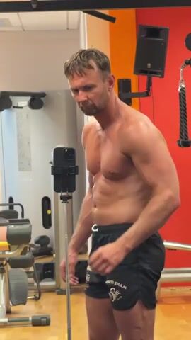 A man with short brown hair, wearing black athletic shorts, stands shirtless in a gym, posing for a video. He is positioned in front of a mirror, with a smartphone on a tripod in front of him. Behind him, gym equipment is visible, including weight machines and red walls. The man flexes his muscles, showing his chest, arms, and abs. The video captures a series of poses, with the man turning slightly to showcase his physique.