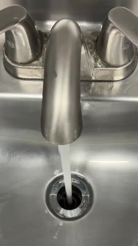 A stainless steel sink faucet dispenses a stream of running water into the drain. The faucet has two handles, one on each side, and a central spout. The sink basin is also made of stainless steel and shows some signs of water spots and minor discoloration around the drain. The water flows directly down the drain.