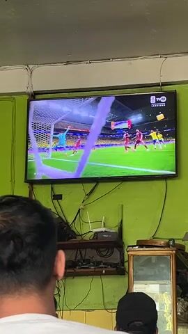The video shows a large television screen displaying a soccer match. The game is in progress, with players in red and yellow uniforms visible on the field. The camera is positioned behind two people who are watching the game. The television is mounted on a green wall, with various electronic devices and cables visible underneath it. There is also a small mirror on the right side of the frame. The overall setting appears to be an indoor space, possibly a shop or a home. The broadcast on the TV shows a close-up of the goal area, with a player in a red jersey attempting to score while a goalkeeper in a yellow jersey defends. The crowd in the background is blurred but appears to be wearing yellow. The top right corner of the screen displays a TV channel logo and what appears to be a match indicator.