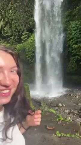 Two women are standing in front of a tall waterfall. The woman on the left is wearing a white t-shirt and a gray plaid dress. The woman on the right is wearing a pink and white striped sweatshirt and a pink baseball cap. They are holding hands and have their arms raised in the air, pointing towards the waterfall. The waterfall is surrounded by lush green vegetation. The ground in front of the waterfall is rocky and wet.