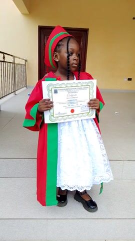 The video shows a young Black girl wearing a graduation gown and cap, holding a certificate. She is standing in front of a building, with a tan wall and a wooden door behind her. The girl has braided hair and is smiling at the camera. She is wearing a red gown with green trim and a white dress underneath. The certificate is white with a green border and appears to be from "KINGDOM HERITAGE MODEL SCHOOL, AKANPAI." The girl is holding the certificate with both hands. The video is shaky and appears to be filmed from a low angle. There is a railing in the foreground. In the last frame, a hand is seen adjusting the girl's cap.
