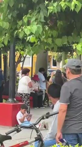 The video depicts a public square filled with people on what appears to be a sunny day. There's a young boy sitting on the ground, focused on a smartphone. A group of adults stands nearby, some conversing and others observing something out of frame. A bicycle is parked in the foreground, partially obstructing the view. The square has trees, park benches, and some buildings with a yellow facade in the background. Music is playing in the background.