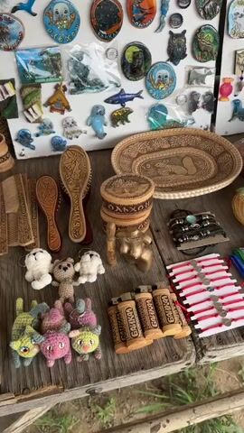 The video pans across a wooden table displaying a variety of handcrafted items and souvenirs. The items include wooden combs and brushes, small crocheted animals, various bracelets made of beads and leather, carved wooden trinkets and boxes, painted stones featuring animal figures, and decorative magnets and ornaments depicting seals, fish, birds, and landscapes. There are also some metal mugs and wooden bottles visible. The overall setting appears to be an outdoor market stall. The camera movement is slow and steady, providing a detailed view of the merchandise. No people are visible interacting with the items.