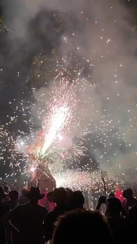 The video shows a nighttime outdoor event with fireworks and a crowd of people. The fireworks are colorful and bright, creating a spectacular display of light and sparks. The crowd is silhouetted against the fireworks, and some people are holding up their phones to record the event. The overall atmosphere appears festive and celebratory.