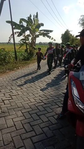 The video shows a group of young people in military-style uniforms, some with bibs bearing numbers. They are standing in formation on a paved pathway. The pathway is made of interlocking bricks and is lined with banana trees and greenery on the left side. In the background, there is a clear sky and some distant trees. A red motorcycle is parked to the right of the formation, and its front wheel and part of its body are visible. The individuals appear to be participating in an organized event, possibly a competition or a parade, given the bib numbers. The lighting suggests it is daytime, likely morning or afternoon, with strong sunlight casting shadows on the ground.