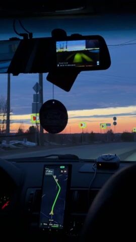 From the interior of a car driving along a rural road at dusk, the sky shows a palette of blues, oranges, and pinks. The dashboard features a navigation display showing a green route, a radar detector, and a rear-view mirror that also functions as a dashcam. A Ford-branded air freshener dangles from the rear-view mirror. Road signs indicating turns are visible outside. The camera is static, offering a driver's view of the road and surrounding landscape.