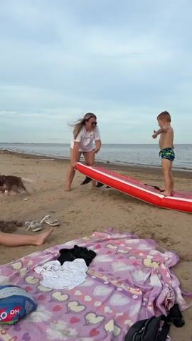 In this video, a woman and a young boy are on a sandy beach near the water. The woman, wearing sunglasses and a white t-shirt with a graphic print, is holding an inflatable red paddleboard. The boy, in patterned swim trunks, stands on the paddleboard. The scene appears to be a recreational outing by the sea under an overcast sky. The time of day is likely late afternoon or early evening, given the soft lighting. The general atmosphere seems relaxed and playful, as they prepare to engage in water activities. A patterned blanket with heart designs, a pair of sneakers, and a blue and white inflatable toy are visible on the sand, suggesting a family outing. The water is calm, with a distant boat visible on the horizon.