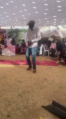 Here's a detailed description of the video:

The video shows a man in a light-colored, long-sleeved shirt, blue jeans, and a dark cap standing in an open, tented area. He gestures with his hands as if speaking or giving instructions to a group of people. Several children and adults are seated or standing in the background, watching him.

A table decorated with a pink cloth and a gift basket is visible to the left of the man. A red carpet is spread out in front of him on the ground. The tent is covered with a white fabric with polka-dot designs. The ground appears to be covered with grass and some loose debris.