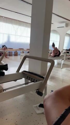 Here is a description of the video.
The video shows a pilates studio with three women participating in reformer exercises.  The first woman in the foreground, dressed in a dark top and shorts with green socks, is working on the reformer. Her foot is strapped to the machine, and she is moving the carriage back and forth, guided by an instructor.  The second woman in the middle of the room is on her hands and knees, stretching or preparing for an exercise. The third woman in the background, dressed in a black sports bra and leggings, is also working on a reformer, facing away from the camera. The studio is bright with natural light coming through large windows covered with sheer white curtains. There are several reformers lined up in the room, as well as wooden tables and chairs. The walls are painted white, and the overall environment is clean and modern.