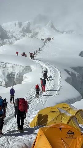 The video shows a mountainous, snowy landscape. In the foreground, several yellow tents are pitched in the snow. Beyond the tents, a winding path of compacted snow leads uphill, marked by footprints. Numerous people, dressed in winter climbing gear and carrying backpacks, are following this path. They are moving in single file, spaced apart from each other. Some individuals are closer to the camera, while others are further up the trail. The background is dominated by snow-covered peaks and misty, cloudy skies. The overall impression is of a climbing expedition on a large, snow-covered mountain.