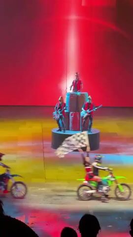 The video shows a performance with motorcyclists and a band on a stage. The stage has a yellow and orange floor, a red backdrop that changes to black, and some pixelated designs. In the center, a band consisting of a drummer on top of a stack of barrels, a guitarist and a keyboardist are playing. The motorcyclists, wearing colorful outfits and helmets, ride around the stage waving checkered flags. The video follows the action, sometimes zooming in on the motorcyclists.