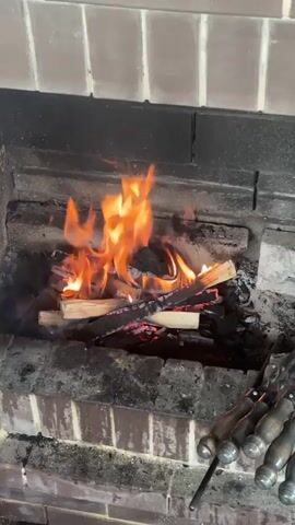 The video shows a close-up view of a fireplace with a fire burning. Several logs are arranged in the center of the fireplace, with flames rising from them. The fire is producing smoke, which drifts upwards. The interior of the fireplace is made of brick, and there is a hearth area at the bottom. In the foreground, on the right side of the frame, a set of fireplace tools, possibly poker and tongs, are visible. The lighting is warm, emanating from the fire. The camera appears to be positioned at eye level, looking into the fireplace. The overall scene suggests a cozy and inviting atmosphere.