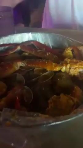 The scene is set in a restaurant with dim lighting. A large metal serving tray filled with seafood sits on a table. The tray contains crab legs, clams, and what appears to be fried food, all covered in a sauce. The tray is lined with a clear plastic bag. A man wearing a white t-shirt is seated at the table behind the tray. He's holding a small white object in his hands. A glass of dark liquid and a bottle of water are also on the table. In the background, there are red chairs and bar stools.